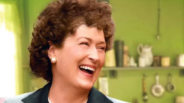 Meryl Streep dressed as Julia Child laughs in a green kitchen.
