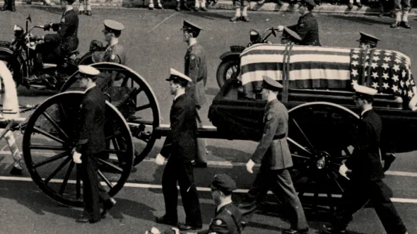 A black and white photo of military men marching beside a coffin covered in an American flag on a flatbed wagon.