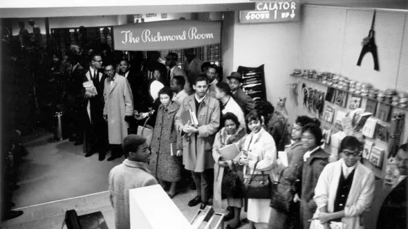 A black and white photograph of people in 1960s attire standing in line inside a department store