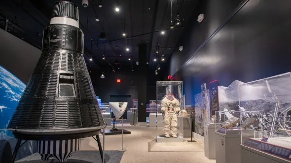 An interior of a gallery with a space capsule, photo of Earth and surface of the moon, and a spacesuit in a display case.
