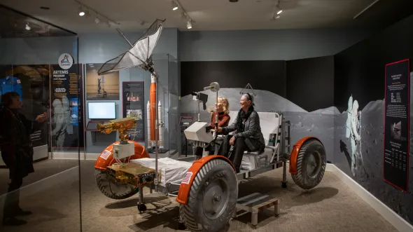 Two people sit in a model moon rover