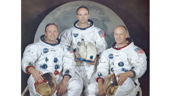 Apollo 11 crew: 3 astronauts in spacesuits hold their helmets and pose in front of a large photograph of the moon