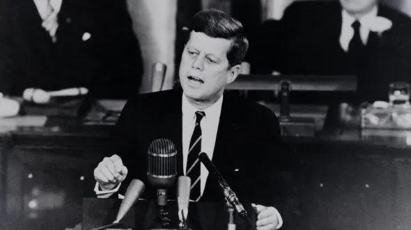Black and white photo of John F. Kennedy Jr. speaking at a podium in front of a large microphone