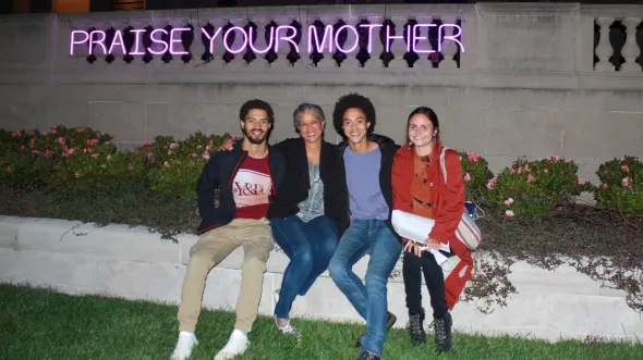 4 smiling people sit on a low stone wall in front of purple neon letters that read "Praise Your Mother"