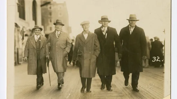 A sepia photograph of 5 men in long coats and hats walking a boardwalk
