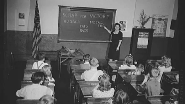 A black and white photo of a classroom. The words SCRAP FOR VICTORY, RUBBER, METAL, FATS, RAGS are written on the chalkboard.