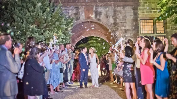 Bride and groom leaving reception walking under brick archway with guests lining path