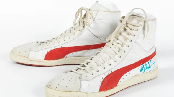 A pair of large white hightop sneakers with a red stripe along the bottom. The signature of Ralph Sampson in green marker is on the lower right heel