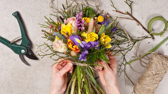 History Blooms Two hands tie ribbon around a bouquet of flowers. Twine and plant sheers are on the table.