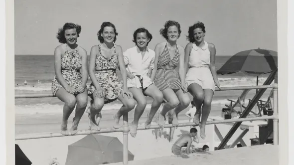 A row of women in 1950s-era dresses and bathing suits sit on a metal railing in front of an ocean beach.