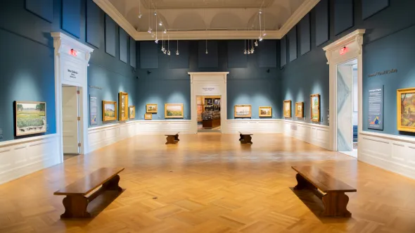 A gallery with blue walls, parquet floors, and landscape paintings on the walls.