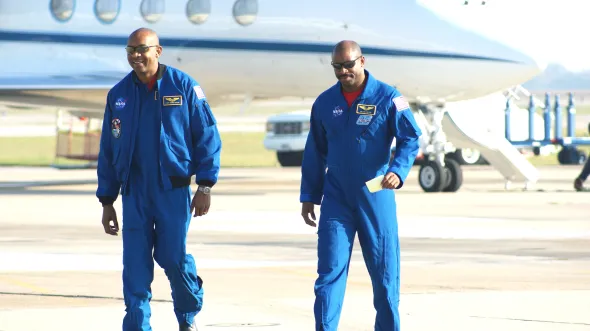 Leland Melvin and Robert Satcher wear blue astronaut uniforms and walk on an airfield with a stationed plane behind them