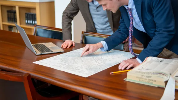 Two people lean over a library table pointing at a map, next to a laptop screen, pencil, and large open book.