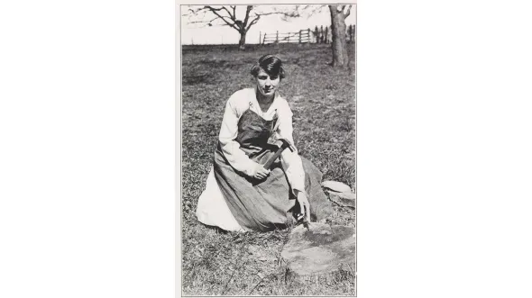 A black and white photo of a woman kneeling in a field wearing a prairie dress and apron