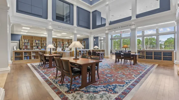 A double-height research library with lamps on research tables, a patterned rug, book stacks, and a wall of windows.