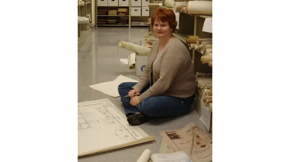 A woman is seated on a floor surrounded by rolled and flattened architectural drawings