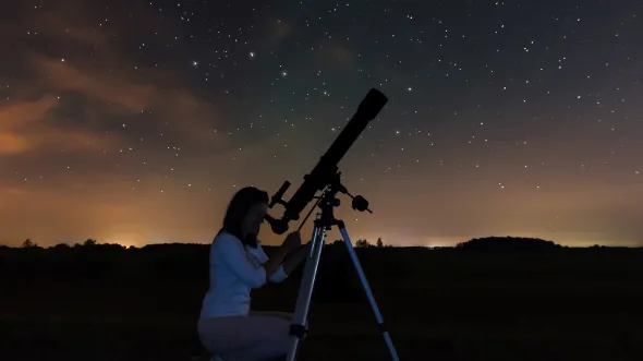 A woman looks through a telescope with a starry sky behind her