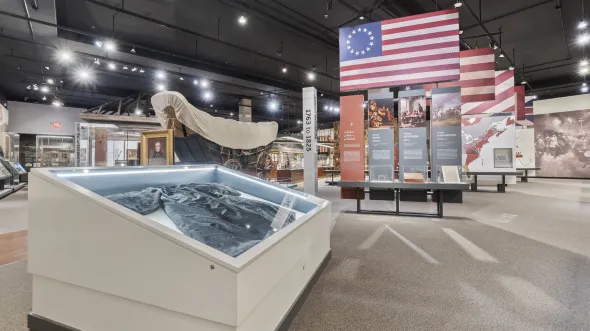 A large display case with a judge's robe in the foreground, with a Conestoga wagon in the background, and historic American flags hang from the ceiling