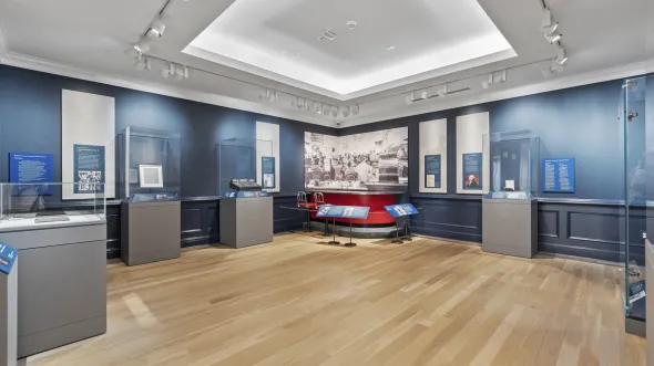 Interior scene of a gallery with wooden floors, blue painted walls, and display cases for artifacts, including George Washington's Diary and a red Woolworth's Lunch Counter