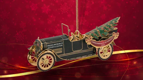 A 3-D ornament of an old open-topped automobile with gold wheels and a Christmas tree in the backseat includes a red ribbon banner that reads Virginia Museum of History & Culture, Commonwealth of Virginia 2022. The ornament is offset by a red background with snowflake and gold ribbon