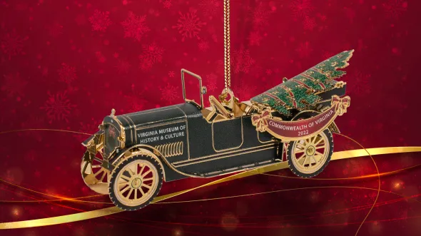 2022 Official Virginia History Ornament A 3-D ornament of an old open-topped automobile with gold wheels and a Christmas tree in the backseat includes a red ribbon banner that reads Virginia Museum of History & Culture, Commonwealth of Virginia 2022. The ornament is offset by a red background with snowflake and gold ribbon