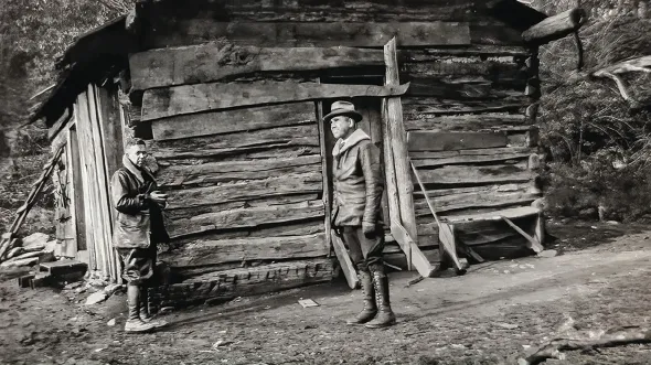 A black and white photo of two people in front of a log cabin
