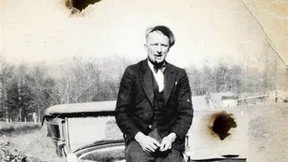 A black and white photo of a man sitting on a car hood.