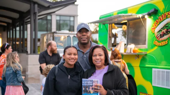 A smiling family of three stand in front of a food truck outside of the museum holding a printed museum scavenger hunt