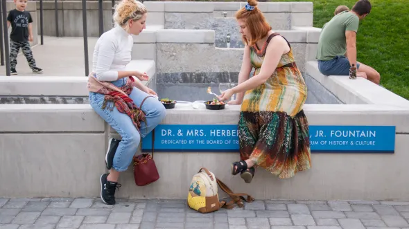 Two people sit on the edge of a fountain eating dinner