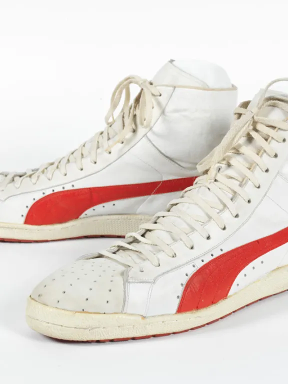 A pair of large white hightop sneakers with a red stripe along the bottom. The signature of Ralph Sampson in green marker is on the lower right heel