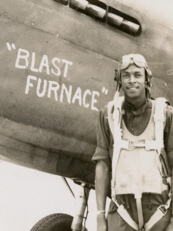 Pilot Clemenceau Givings stands in front of a propeller plane with the words "Blast Furnace" painted on the side