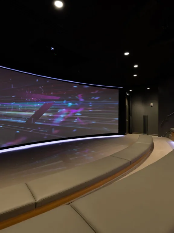 A large curved screen fills one wall of a theater with rows of stadium-style bench seating filling the rest of the space