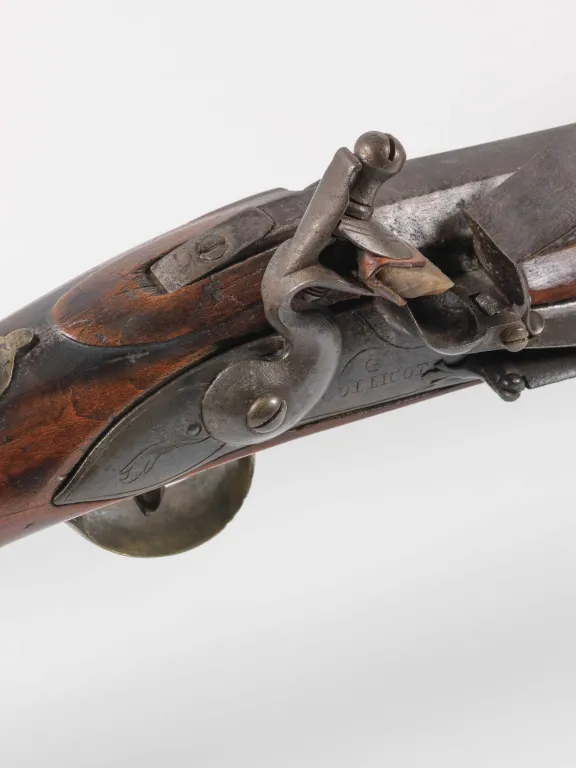 Close-up of a musket's flintlock, with an exterior bridle and is engraved with a floral pattern and is marked "Collicot" 