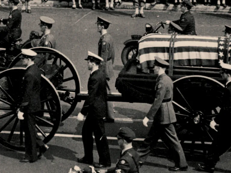 A black and white photo of military men marching beside a coffin covered in an American flag on a flatbed wagon.
