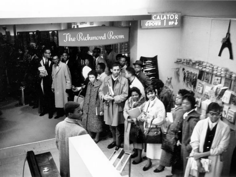 A black and white photograph of people in 1960s attire standing in line inside a department store