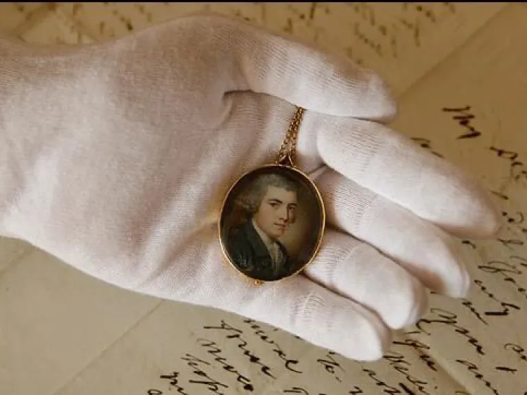 A gloved hand holding a miniature painting over handwritten letters