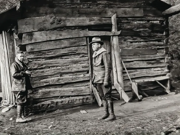 A black and white photo of two people in front of a log cabin