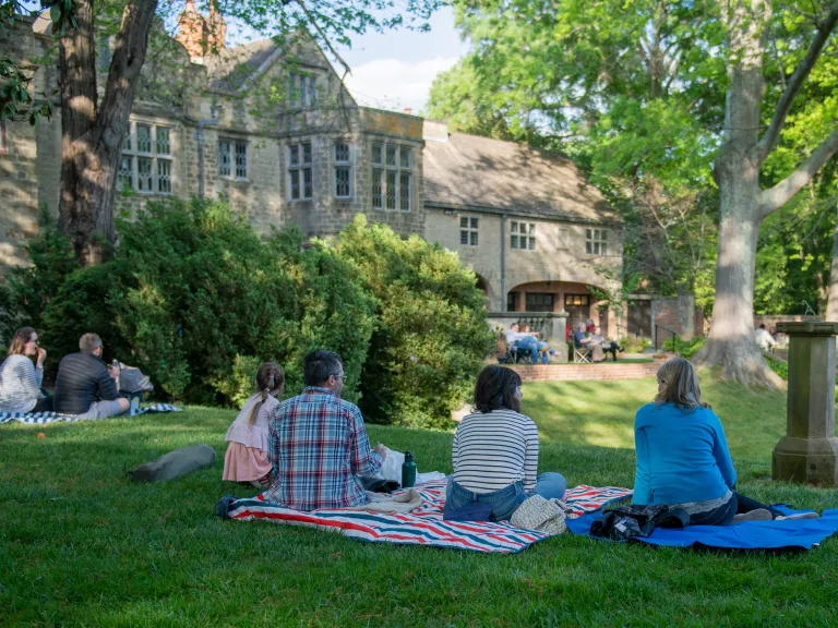 People sit on picnic blankets on a grassy hillside facing a stone Tudor mansion