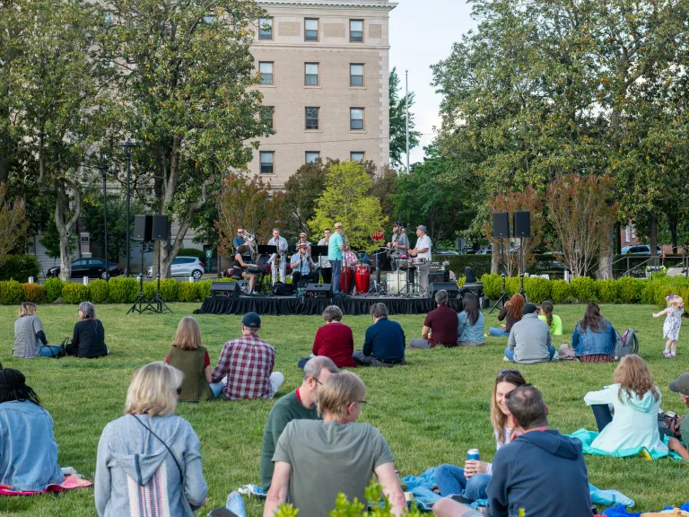 People sit on picnic blankets on a grassy lawn watching a band on a small stage