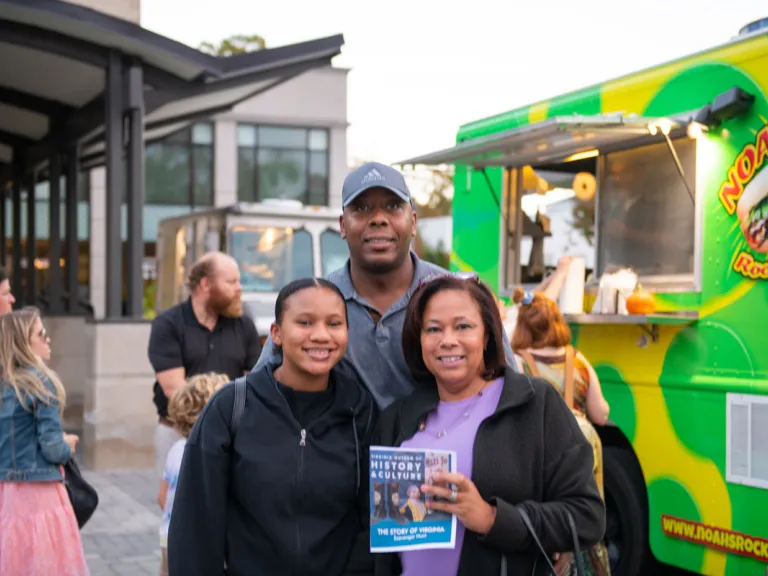 A smiling family of three stand in front of a food truck outside of the museum holding a printed museum scavenger hunt