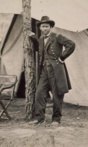 Black and white photograph of Ulysses S. Grant leaning against a tree in front of a tent in a military uniform. 
