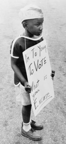  Black and White Photograph of a young African American boy holding a sign saying, "I'm To(o) Young To Vote - What is your Excuse?" 