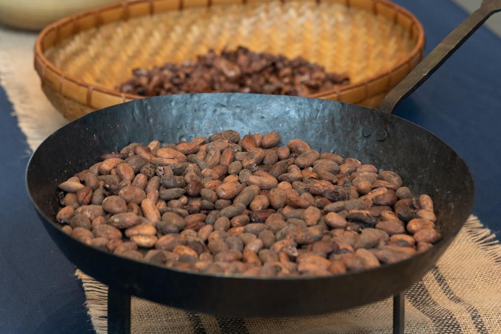 Cocoa beans in a cast iron pan in the foreground, with more beans in a basket in the background